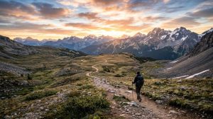 Wide scenic landscape with a lone walker on a long trail stretching toward mountains, sunrise lighting, sense of journey and endurance
